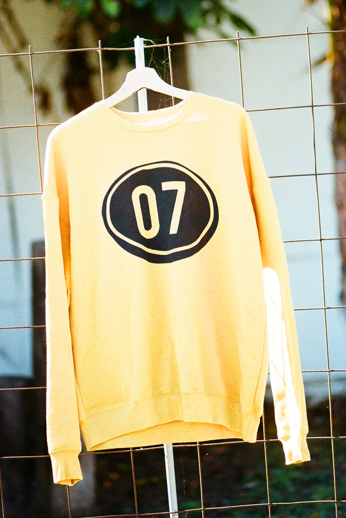 Yellow sweatshirt with a black circle and number 07 on a hanger against a wire fence background.