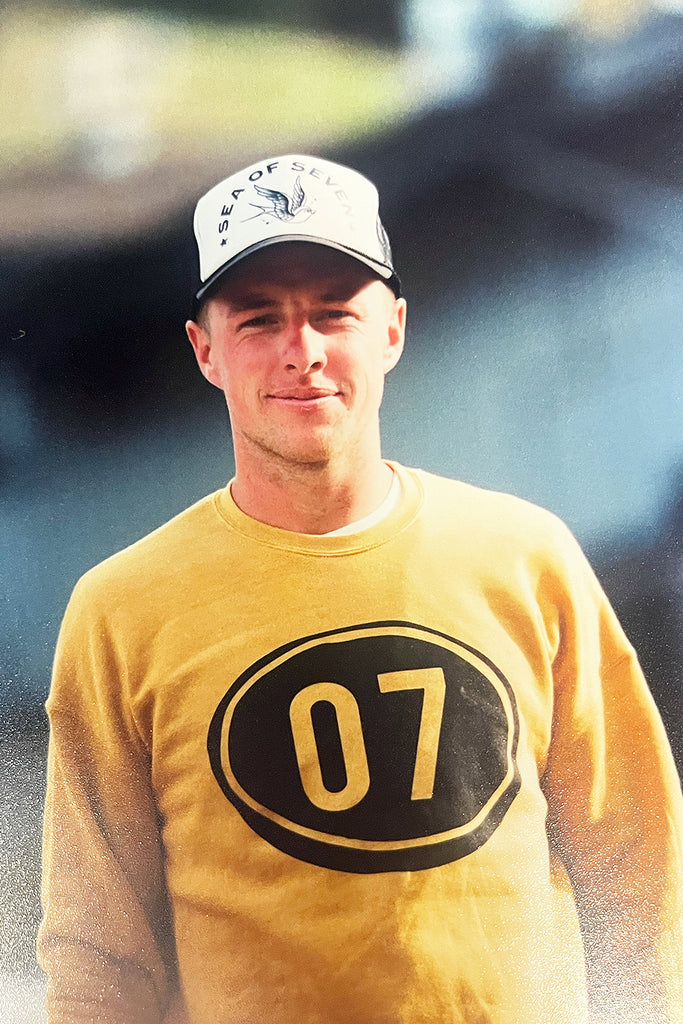 White male wearing mustard and gold sweatshirt with the number 07 and a black and white trucker hat.