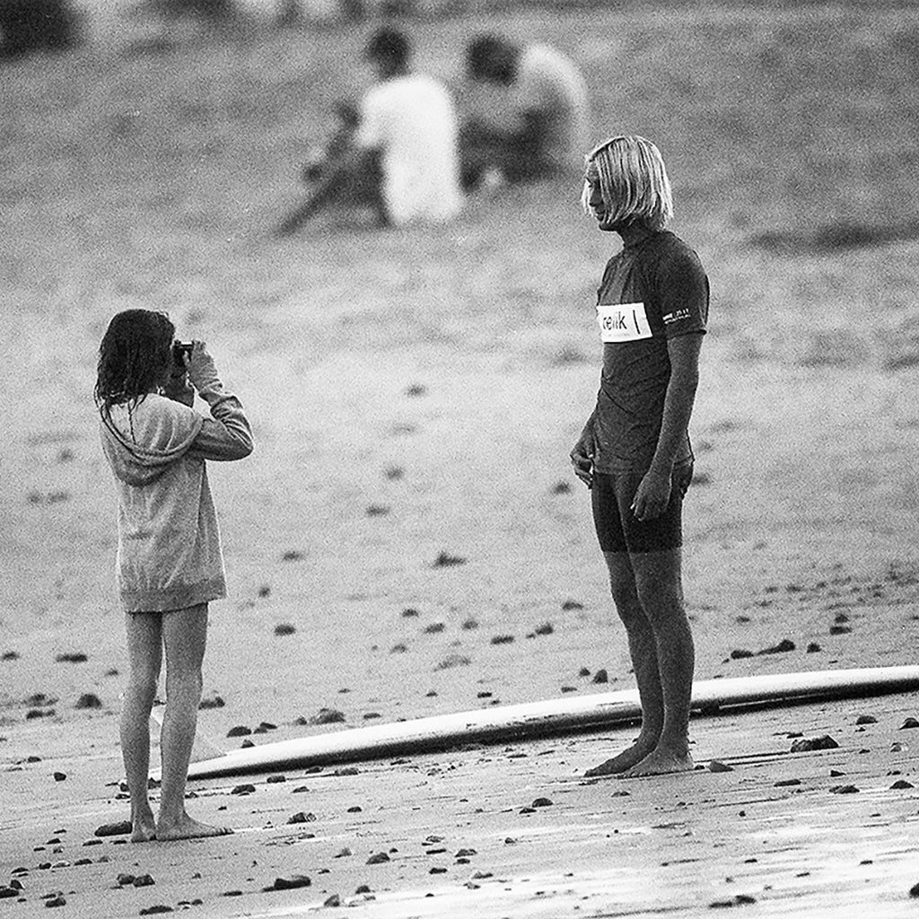 a young girl taking a picture of a surfer on the beach in malibu.