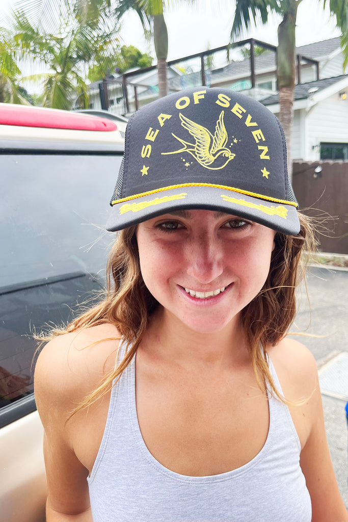 girl wearing a black and gold trucker hat with the words SEA OF SEVEN and a yellow swallow bird drawing.