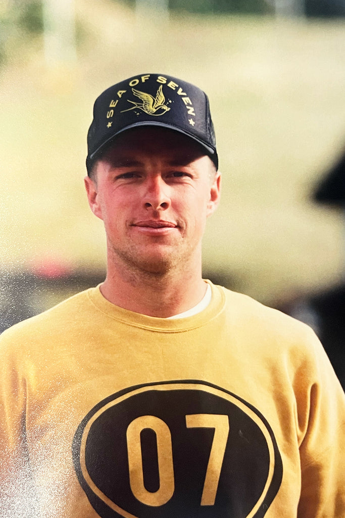 Adult male wearing a black trucker hat with the words sea of seven and a swallow bird graphic and a yellow mustard sweater with the numbers zero seven.