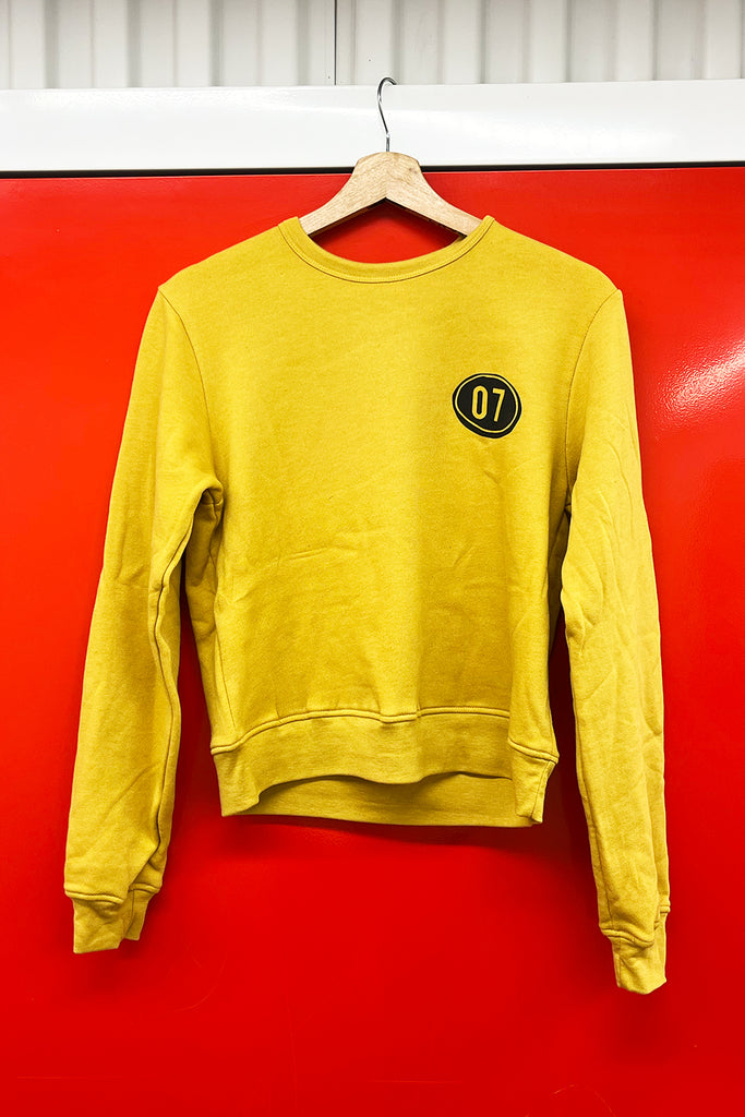 Yellow sweatshirt with a black and gold emblem of a 07 'zero seven' on a hanger against a red wall.