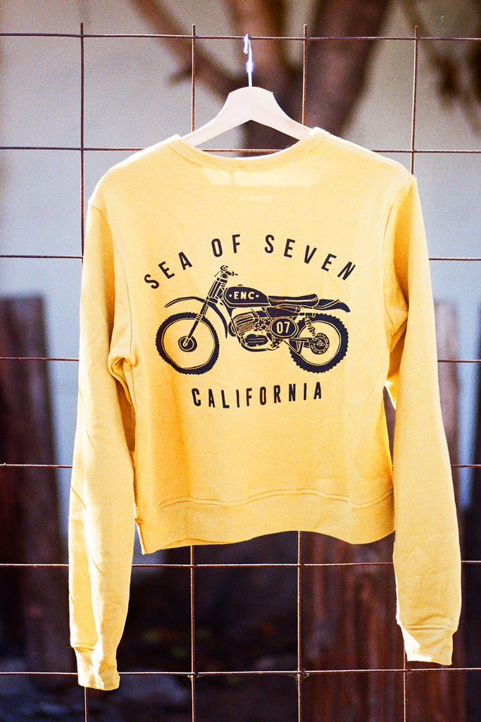 Yellow sweatshirt with motorcycle graphic and text "SEA OF SEVEN CALIFORNIA' on a hanger against a tiled wall.