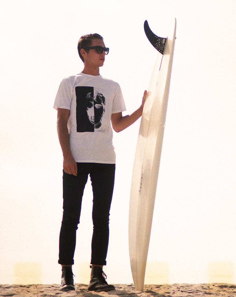 men holding a single fin surfboard on the beach wearing sunglasses, white tee and black pants and boots.