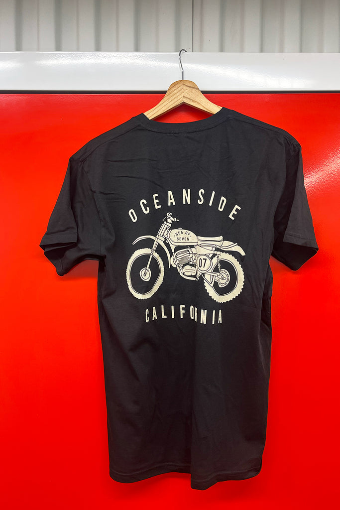 Black t-shirt with a graphic of a vintage motorcycle and text, hanging on a wooden hanger against a red background.