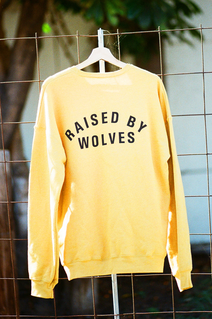 Yellow sweatshirt with 'Raised by Wolves' text on a hanger against a wire fence background