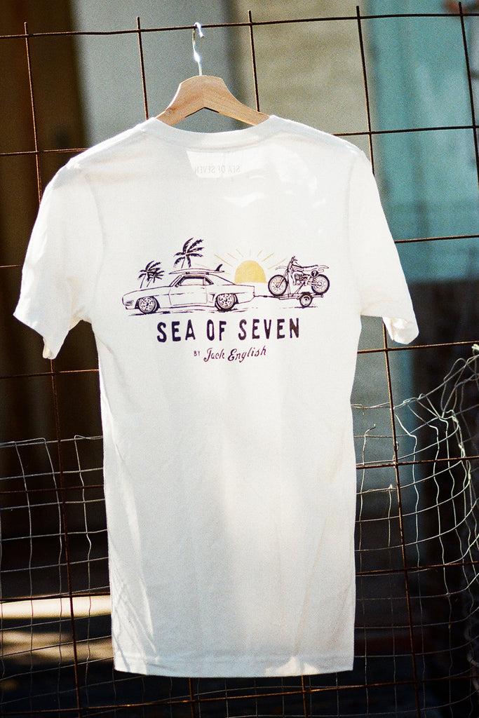 White Sea Of Seven by Jack English tee on hanger with a graphic of a vintage car with a surfboard towing motorcycle dirt bike.