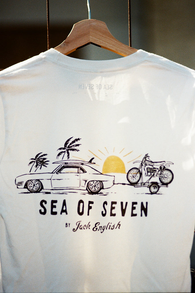 White t-shirt with 'Sea of Seven' graphic design featuring a car, motorcycle, and palm trees on a hanger.