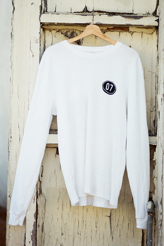 White long-sleeve shirt with a 07 logo on a wooden hanger against a rustic wooden door.